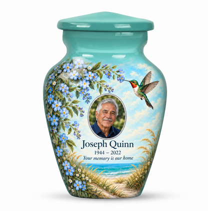 One seafoam green hummingbird keepsake urn with blue forget-me-nots and a coastal scene, Joseph Quinn sample portrait and memorial text on white.