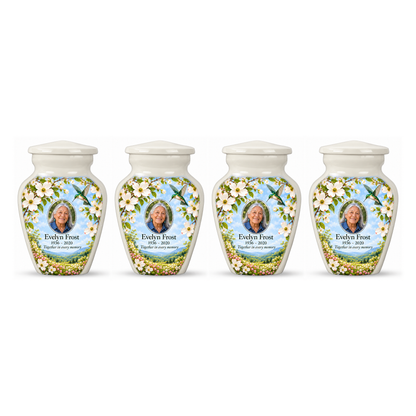 Four white hummingbird keepsake urns in a row on white, each with white dogwood artwork and Evelyn Frost sample portrait and memorial text.