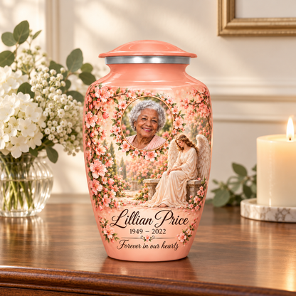 Pink adult urn with a seated angel, peach blossoms and an oval floral sample portrait frame, displayed on a memorial console.